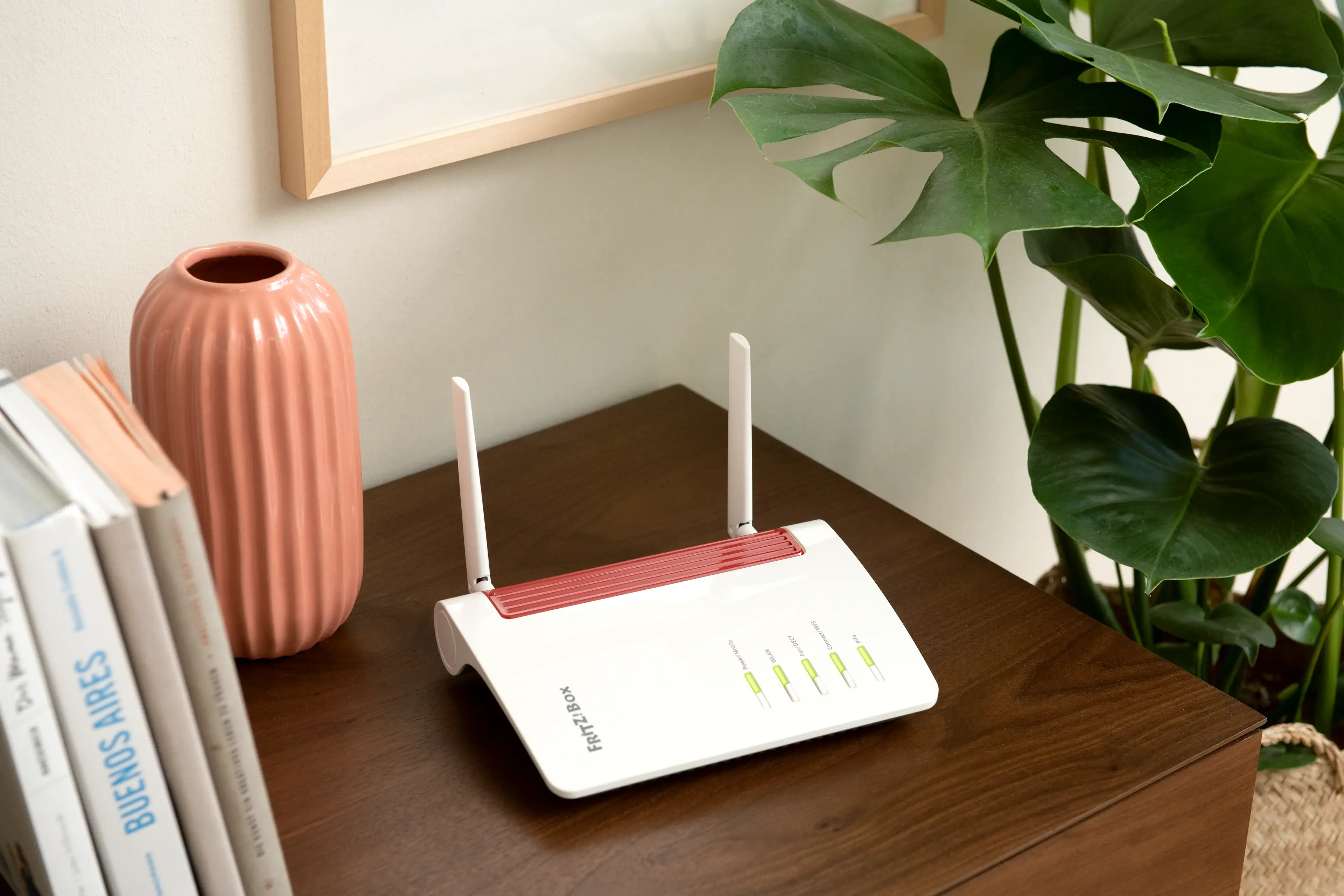 Weißer WLAN-Router mit roten Akzenten auf einem Holztisch, umgeben von Pflanzen und Büchern.